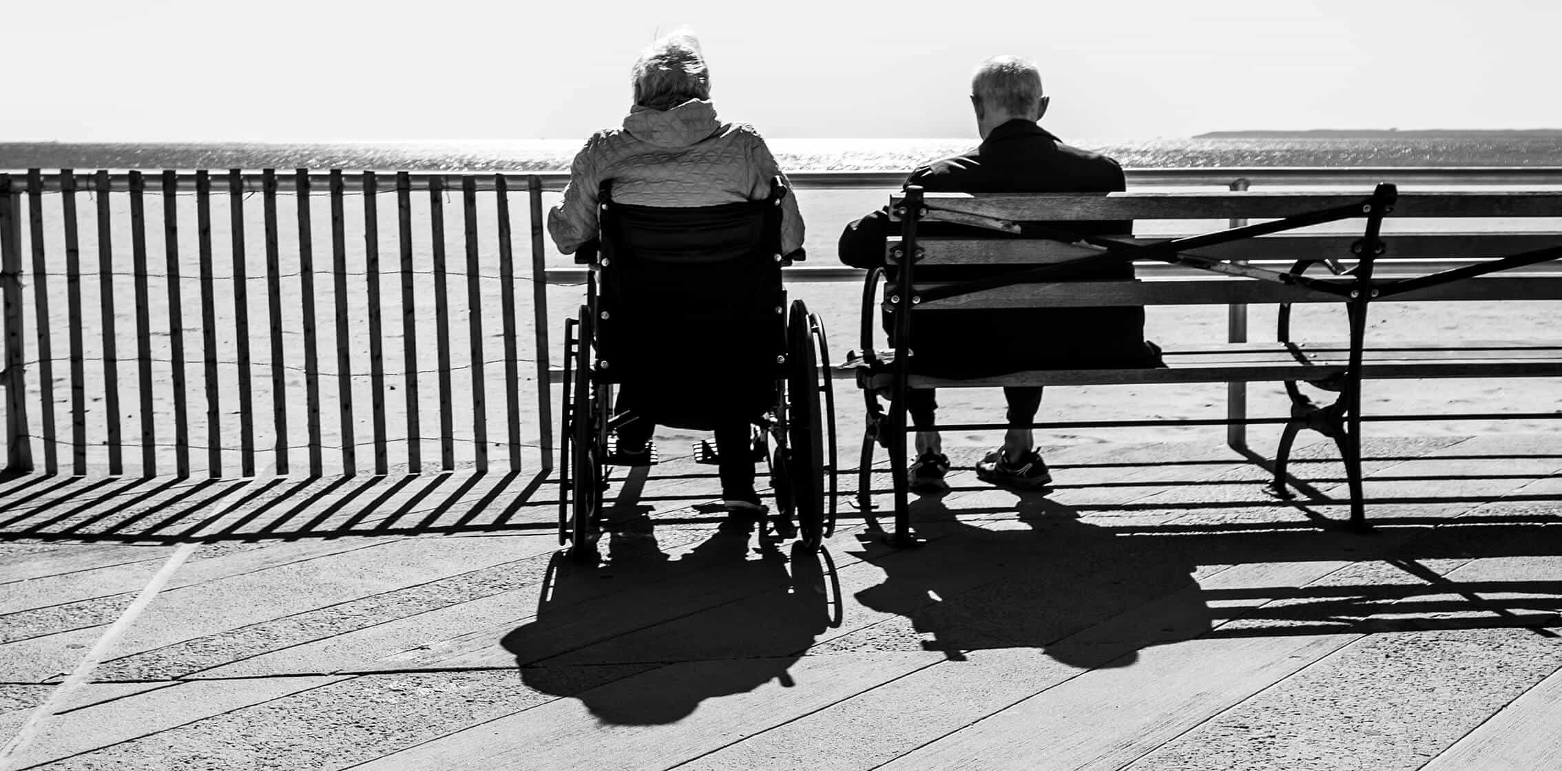 Two people sit by a railing overlooking the sea, one benefiting from custom wheelchair solutions and the other on a bench, shadows gracefully cast on the wooden boardwalk. This striking black and white image captures the serene moment with subtle elegance.